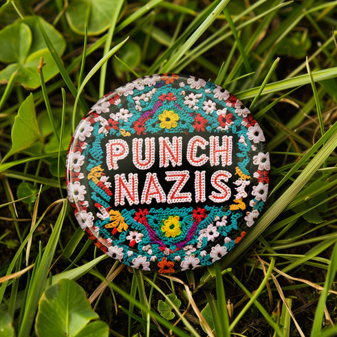 Round pinback metal button with an embroidered-style design on a black background. The center displays the phrase "PUNCH NAZIS" in bold, white stitched-textured lettering with red accents. Surrounding the text is an elaborate folk-inspired floral pattern, densely packed with embroidered-style flowers in shades of red, yellow, blue, pink, and white. The intricate design includes symmetrical lace-like stitching and decorative motifs, creating a rich, tapestry-like effect.